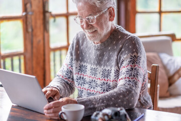 Smiling white haired senior man in winter sweater sitting in living room using laptop computer. Carefree elderly grandfather enjoying tech and social. Rustic chalet in the wood