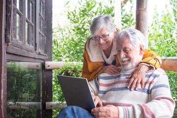 Carefree senior couple hugging in wooden balcony of mountain chalet while man using laptop computer. Caucasian elderly people relaxing outdoor in mountain
