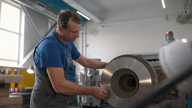 The turner processes the metal part on a lathe in a lathe shop. Slow motion video. Medium shot. A man of 50 years in overalls works near the machine.