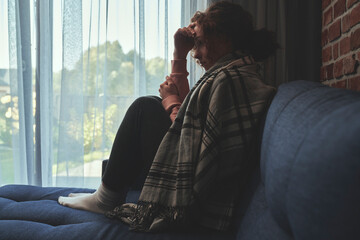 Depressed young caucasian woman covered with blanket on sofa - obrazy, fototapety, plakaty
