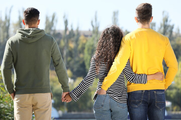 Woman holding hands with another man while hugging her boyfriend outdoors on sunny day, back view....
