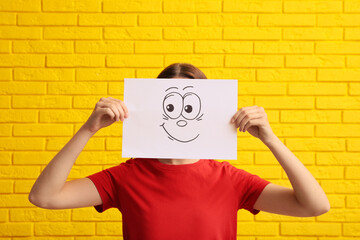 Woman hiding behind sheet of paper with happy face near yellow brick wall