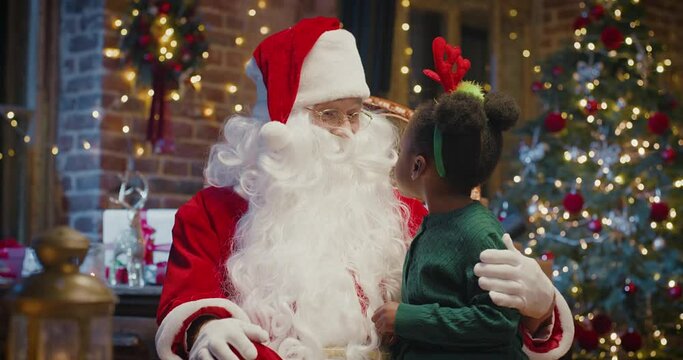 Happy little multiracial girl sitting on Santa Claus knees and telling something to him with fireplace in the background