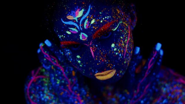 Woman with body art made with luminous fluorescent paints, open eyes and looks into camera, dark background. Female model in studio painted glowing colors