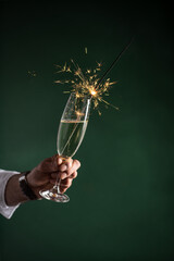 A male hand in white shirt holding glass of sparkling wine champagne with sparkler in it on dark...
