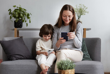 Laughing woman texting typing message on telephone having positive emotion. Smiling male kid playing online internet game use smartphone. Modern family relaxing on couch with digital electronic device