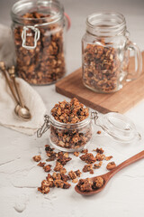 Mason jar with granola breakfast cereal with nuts. wooden spoon with scattered granola in front and two jars with granola on small wooden board in the back on the off-white surface