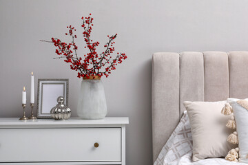 Hawthorn branches with red berries, candles and frame on chest of drawers in bedroom