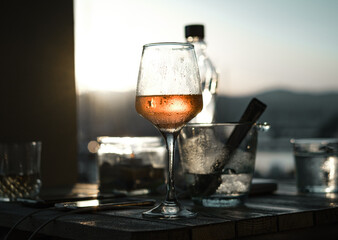 Sunset glass of wine, ice , bottle of water, sun, grey dark tones, against light shot, evening apperitive drink, sun blink in a glass