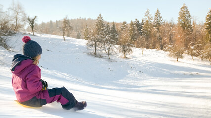 Fototapeta na wymiar Llittle girl sledding with a warm knit hat, laughing descending the icy slope on the yellow shovel sledge.