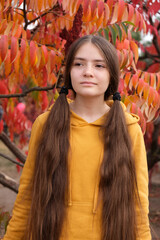 A 13-year-old girl with long hair with two tails in an orange hoody against a background of red leaves.