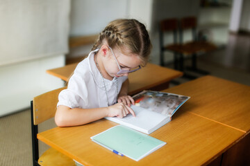cute girl child with glasses reads a book at a desk at school, learning concept, private small school