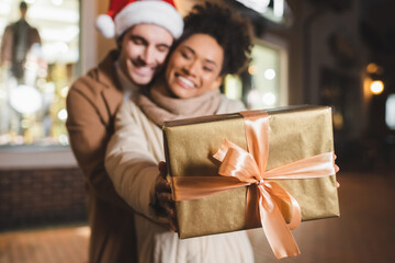 Fototapeta na wymiar blurred and happy african american woman holding gift box near boyfriend in santa hat.
