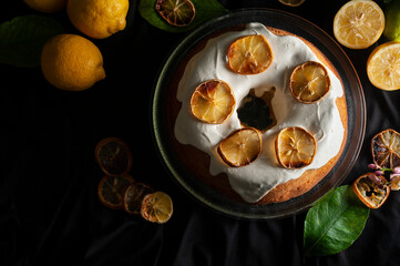 round lemon pudding with cream on top and dried whole and sliced lemons, lemon leaves and flowers with dark background