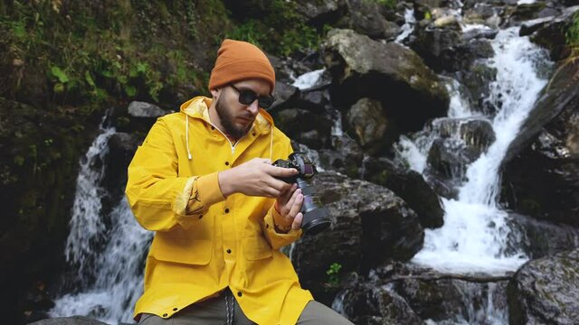 Bearded male tourist in sun glasses with camera takes pictures of mountain river rocky waterfall. Young adult traveler photographer on vacation in highland among rocks. High land natural running water