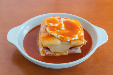 Hot Francesinha sandwich with sausages and meat, melted cheese, poured with beer sauce and topped with a fried egg. Close up in a plate on the table.