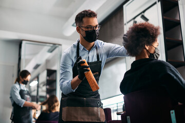 Male hairdresser styles African American woman's hair with hairspray and wears face mask due to coronavirus pandemic.