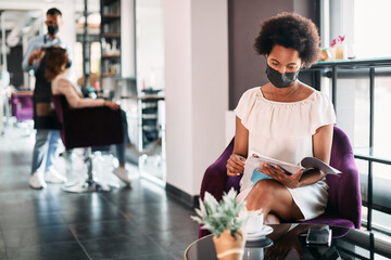 African American woman reads magazine and wears face mask while waiting for hair treatment at...