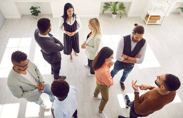 Groups of multi ethnic people standing in office and talking. Diverse men and women communicating during casual corporate meeting, psychological training or after business event. High angle shot - obrazy, fototapety, plakaty