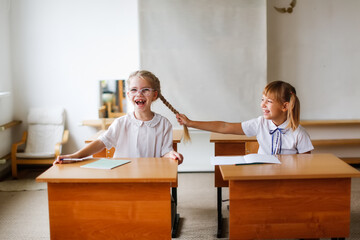 Two schoolchildren, children at their desks in a bright classroom, a girl pulls a classmate by the braid. Classmate relationships and disputes, school learning concept and children's emotions - obrazy, fototapety, plakaty