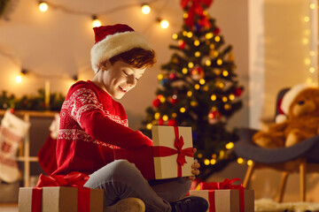 Surprise from Santa Claus. Happy excited boy sits on the floor in a cozy room and opens a Christmas...