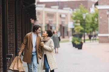 joyful multiethnic couple in coats holding hands while looking at each other and walking with purchases.