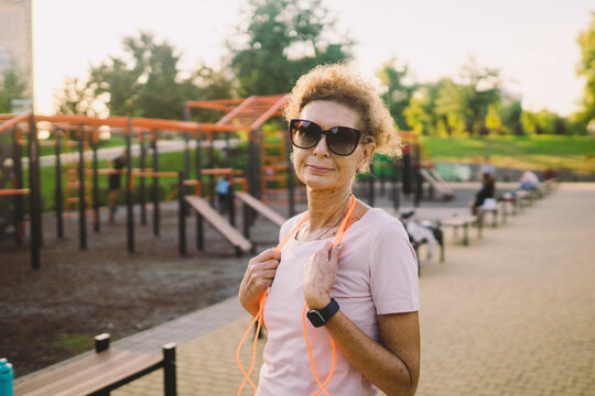 Smiling Beautiful Mature Woman With Skipping Rope Posing Against Backdrop Of Street Gym After Workout. Older Woman With A Jumping Rope Ready For Training. Portrait Of A Sporty Smiling Mature Woman