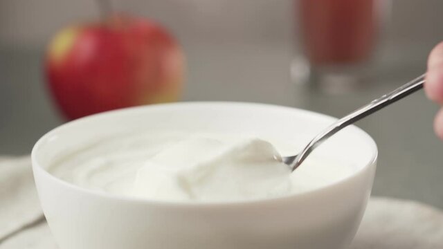 Hand picks up yogurt with a spoon. Bowl of greek yogurt with apple and glass of juice. 
