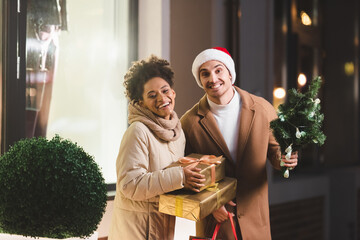 happy man in santa hat holding shopping bags and small fir near african american girlfriend with gift boxes.