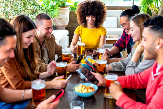 young multiethnic group of friends holding smartphone on social media having fun together drinking happy hour beer at bar restaurant sitting on table indoor. group of diverse people cheering together. - Powered by Adobe