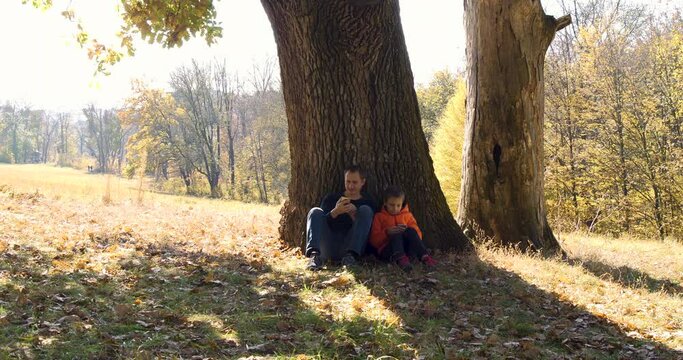 Father And Son Rests Under the Huge Oak in National Park While Hiking in Autumn. Family Use Smartphones in Nature and Talk with Each Other