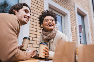 happy interracial couple holding hands and coffee to go near blurred shopping bags.