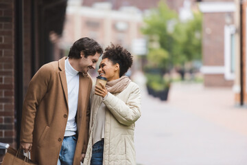 cheerful african american woman holding paper cup near smiling boyfriend with shopping bags.