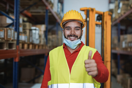 Young hispanic man working inside warehouse while wearing safety face mask under chin and showing thums up on camera