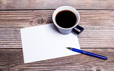 On a wooden table, there is a blue cup with coffee, a pen and a white sheet of paper with a place to insert text. Copy space