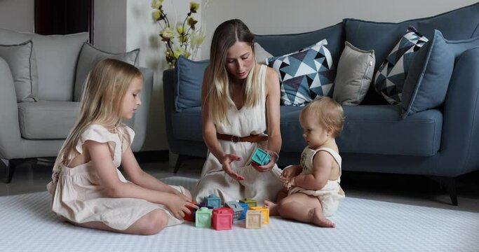 Young caucasian mother or nanny playing with cute baby one year old and elder daughter, learning to build a tower from cubes for kids. Three generation family spending time together and enjoying life