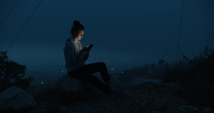 Young millennial woman outdoors during evening dusk scroll through feed on her smartphone. Cinematic screen lit face concept.Generation z and technology. Youth searching online, dating or social media