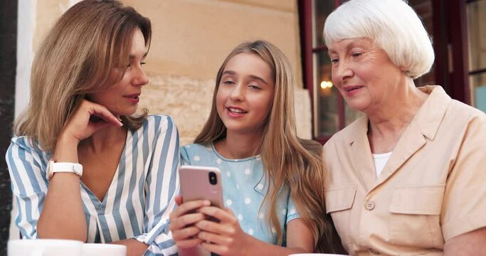 Happy teenage girl sits with her grandmother and mother on terrace of restaurant and shows them interesting news from Internet holding smartphone.