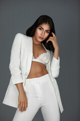 Portrait of an attractive young white woman with wavy black hair and beautiful makeup posing by herself inside a studio with a grey background wearing a white suit with black high heels and white bra.