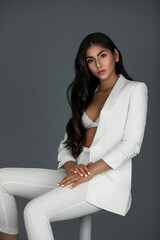 Portrait of an innocent white woman with wavy black hair and beautiful makeup posing by herself inside a studio with a grey background wearing a white suit with black high heels and white bra.