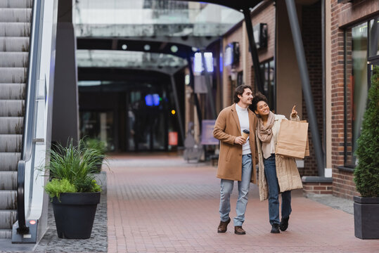 full length of african american woman holding shopping bags and pointing while walking with happy boyfriend in coat in mall.