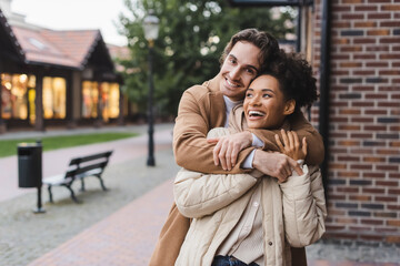 happy man embracing excited african american girlfriend near building outdoors.