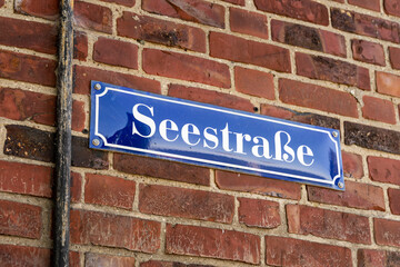 Seestrasse road sign on a red brick wall in Germany. White letters on a blue metal plate. Weathered grungy wall with dirt and cobwebs. Building exterior in a German town.