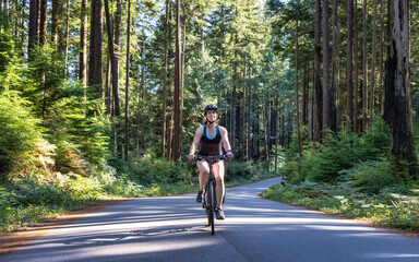 Adventurous White Caucasian Woman on a bicycle riding on a path in Green Canadian Rain Forest. Seymour Valley Trailway in North Vancouver, British Columbia, Canada.