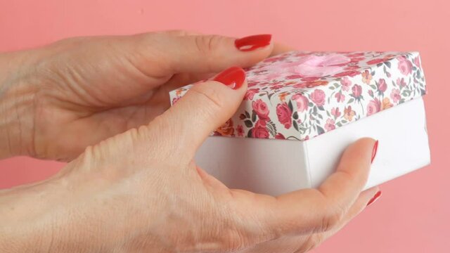 A small gift box with a floral print and a pink bow that is held by the hands of woman whose nails are painted with red nail polish on a pink background