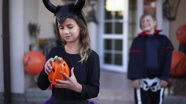 Pretty Caucasian girl in devil costume with horns holding jack o'lantern as boy vampire skeleton laughing standing at background. Cheerful children enjoying Halloween celebration outdoors