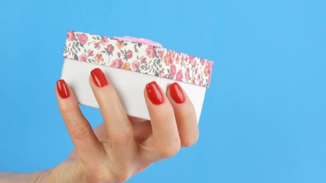 A small gift box with a floral print and a pink bow that is held by the hands of woman whose nails are painted with red nail polish on a blue background