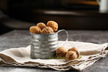A metal mug with whole brown walnuts on a beige colored linen cloth on a grey concrete surface