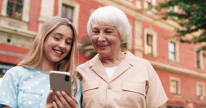 Happy granddaughter and grandmother standing in the middle of city square and laughing looking at funny photos on smartphone.
