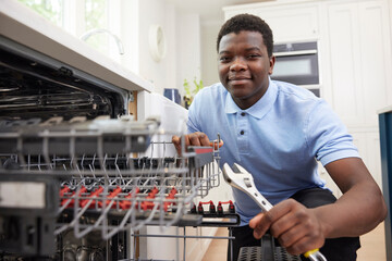 Portrait Of Young Man Training As Plumber Fixing Domestic Dishwasher In Kitchen